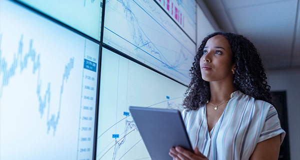 woman analyzing financial charts and data on multiple screens while holding a tablet in a modern trading environment showing stock trends and market analysis for 9 key assets