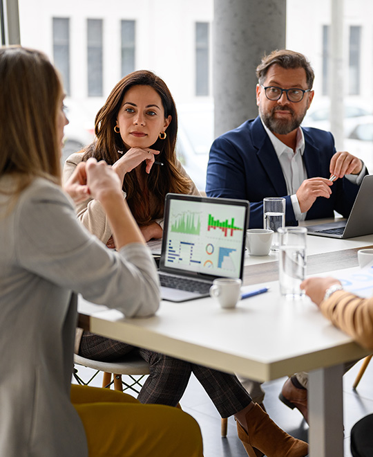 business meeting with five professionals discussing project metrics and charts on laptops at a table